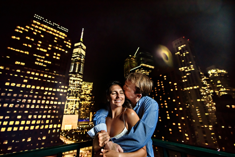 A man kisses his fiance on the rooftop in NYC with the city lights twinkling behind them during their battery park engagement session.